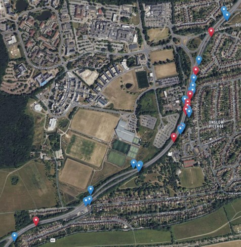 An aerial photograph of a town shows a mix of residential buildings, roads, green spaces, and public facilities. A main road runs vertically along the right-hand side of the image, with numerous red and blue markers positioned along it, indicating NO2 monitoring sites. The area includes housing estates, sports fields, car parks, and other amenities.
Prominent labels on the image include "Onslow Village" situated on the right, and road names such as "Egerton Road" and "A3," helping to identify key routes and neighbourhoods within the town.